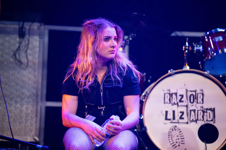 A woman sits down next to a drum kit.