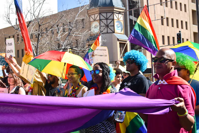 A group of women and men, some in colourful clothing and wigs, carry part of a giant rainbow flag in an urban setting.