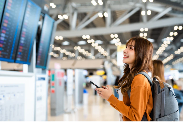 une jeune femme dans un aéroport consulte le tableau des départs, smartphone à la main