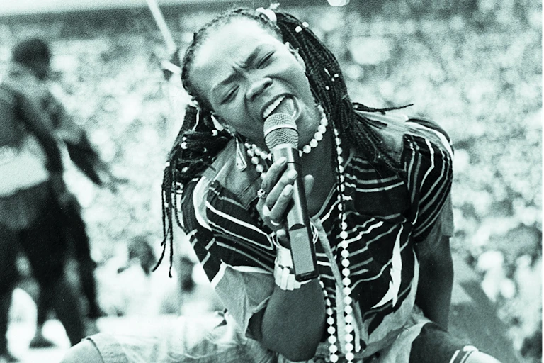 A black and white photo of a woman singinging her heart out into a microphone on stage, her braided hair in pigtails, her eyes shut. She's down on her haunches.