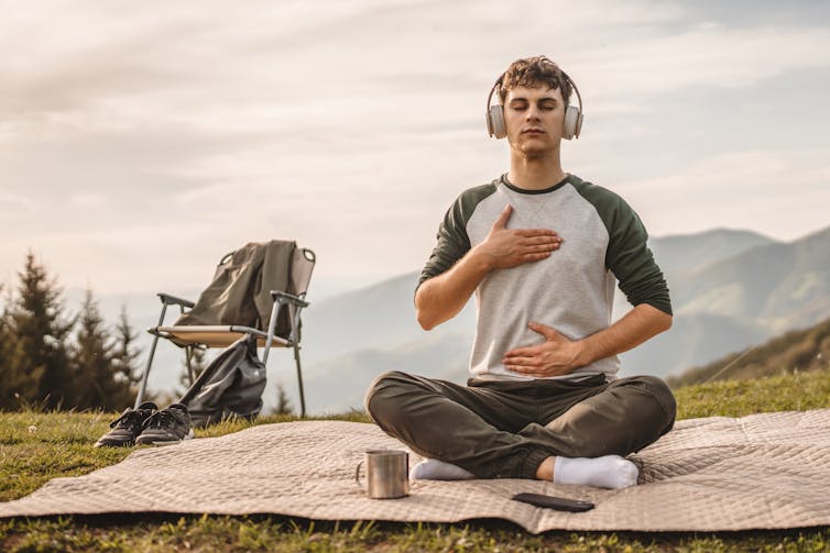 Young man sits on blanket near mountains wearing headphones and meditating