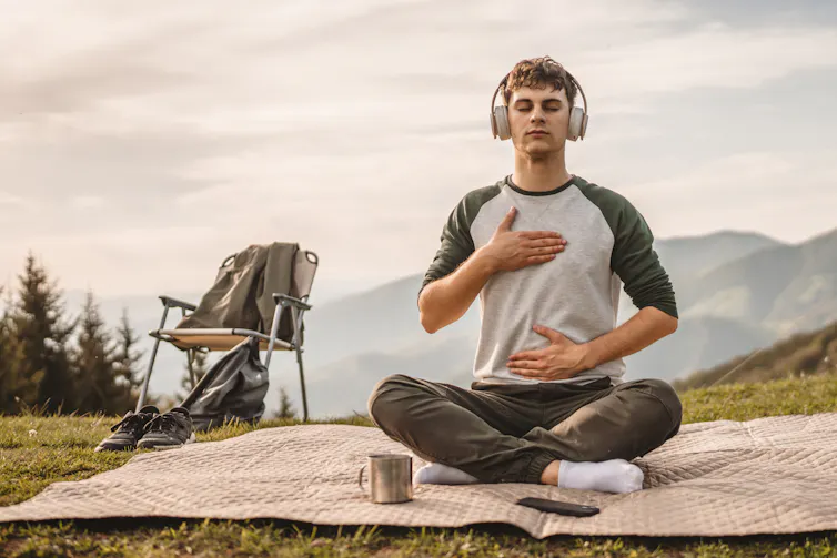 Young man sits on blanket near mountains wearing headphones and meditating