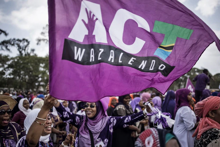 A group of women smiling and cheering while holding up a purple flag that says ACT Wazalendo