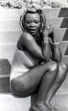 A woman with highlighted braids sits on some stairs in a bathing suit, hands clasped in front of her and a mouth open in exclamation.