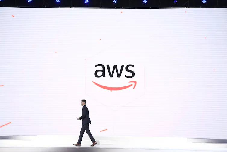 A man walks across a stage in front of a large a w s logo on a white background.