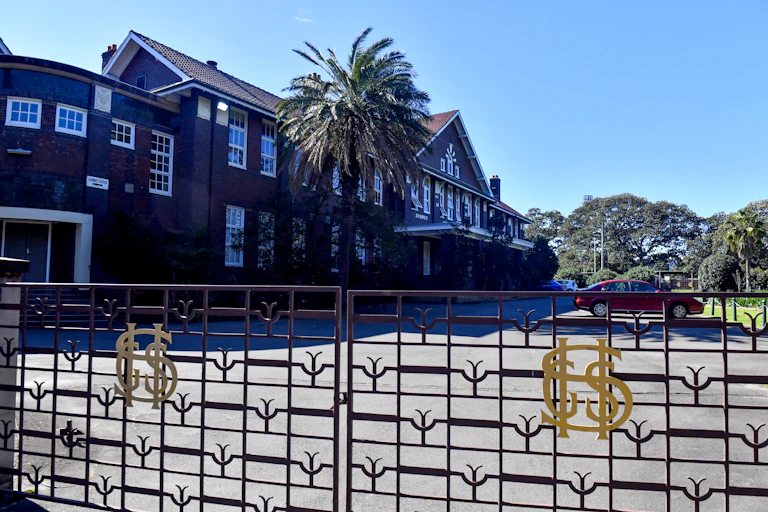 The gates and main building of Sydney Girls High School. It is a two story brick building with car parking at the front.