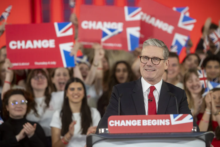 A man in a suit and glasses speaks at a podium while supports hold placards with 'change' on them.