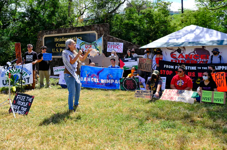 A woman wearing jeans and a keffiyeh, and holding a shell, speaks into a microphone to a gathering of people with Cancel RIMPAC protest signs. Near her feet is a sign saying 'Free the people, free the land'. The sign for the Headquarters of the US Pacific Fleet is visible behind the protestors.