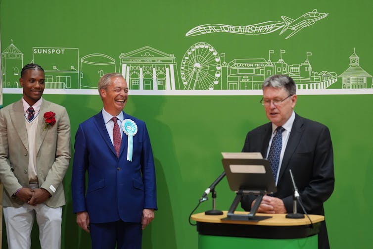 Nigel Farage smiling as a man at a podium speaks.
