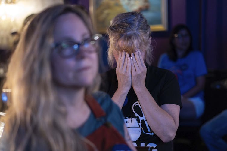 A woman sitting in a room puts her face in her hands, covering her eyes.