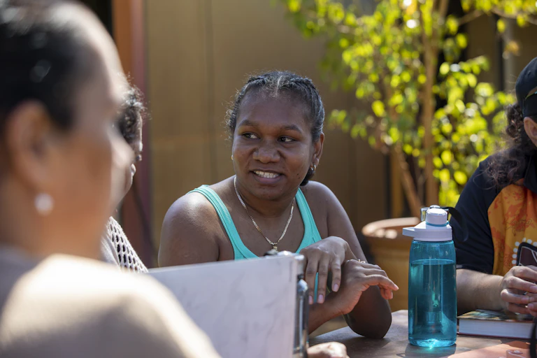 A young woman sits at an outdoor table, talking to other people. There are books and water bottles on the table.