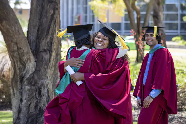 Two people wearing graduating robes hug. One holds a rolled up certificate.