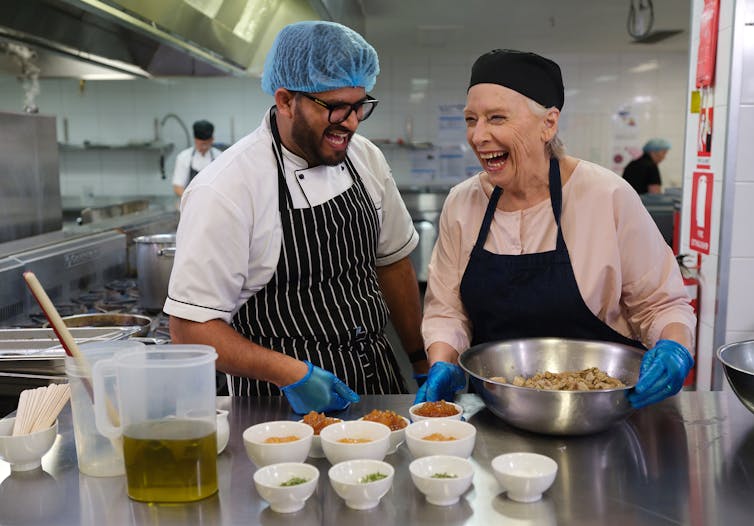 two people stand in commercial kitchen, laughing while preparing food
