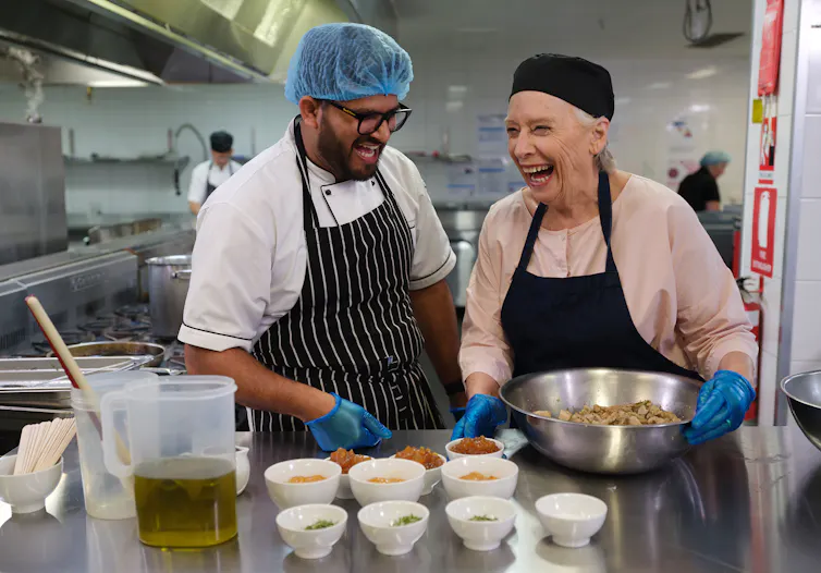 two people stand in commercial kitchen, laughing while preparing food