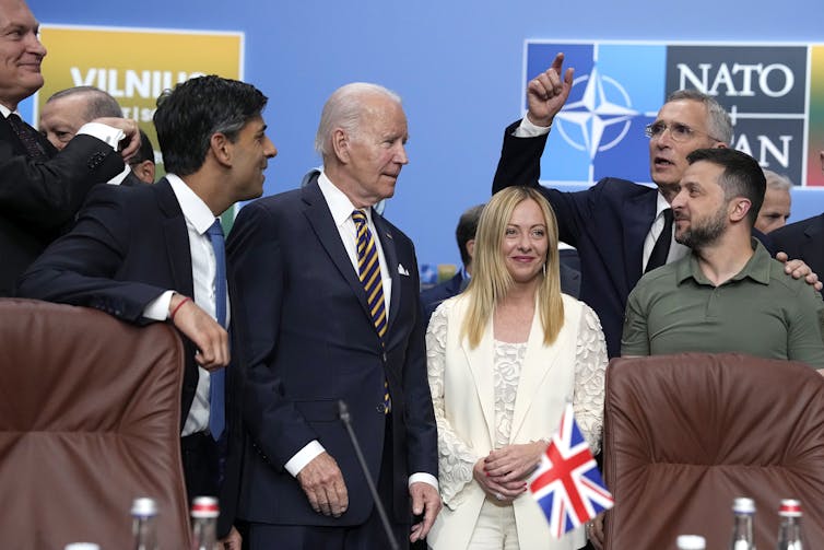 A group of men and women stand chatting in front of a sign reading 'NATO.'