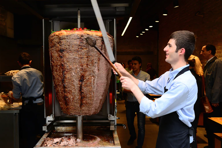 A Turkish man carves meat from a large doner skewer.