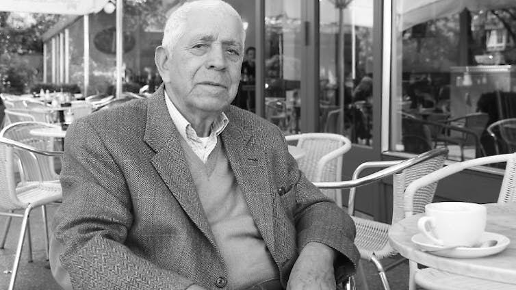 AN elderly man sits at a table at a pavement cafe with a coffee cup in front of him