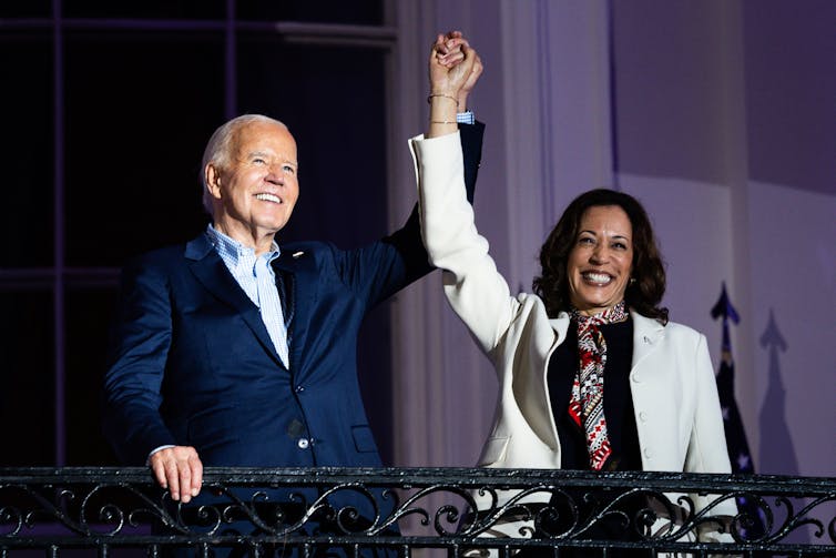 Biden and Harris stood on a balcony holding each other's hand in the air and smiling.