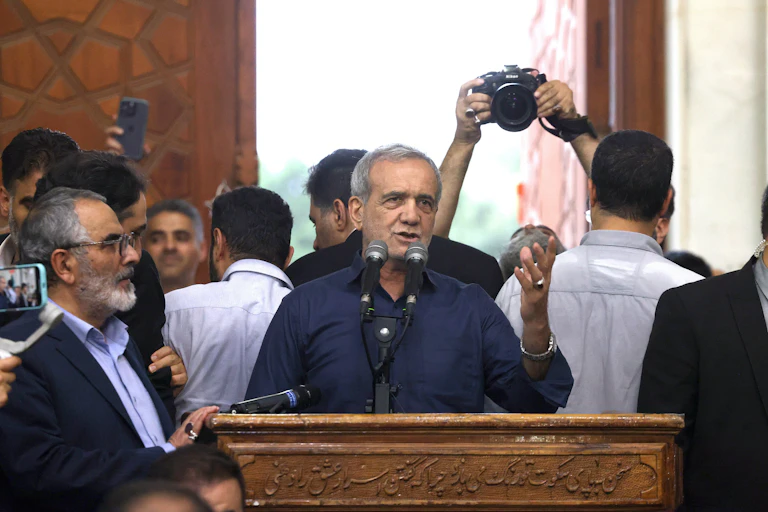 Iranian president-elect Masoud Pezeshkian speaks at a podium, surrounded by supporters. Over his shoulder a phographer holds up a camera.