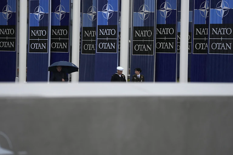 Two military personnel stand beneath a blue NATO banner near someone under an umbrella.