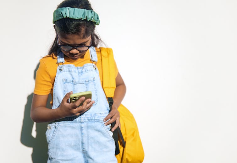 A young girl stands against a white wall, clutching her backpack while engrossed in her phone