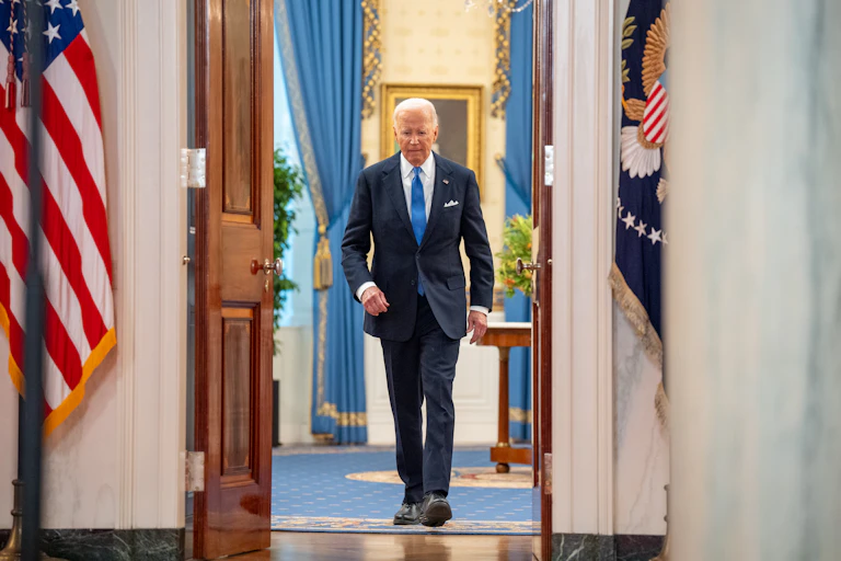 A white man with white hair, a navy suit and a blue tie walks through open doors, with an American flag on one side of it.