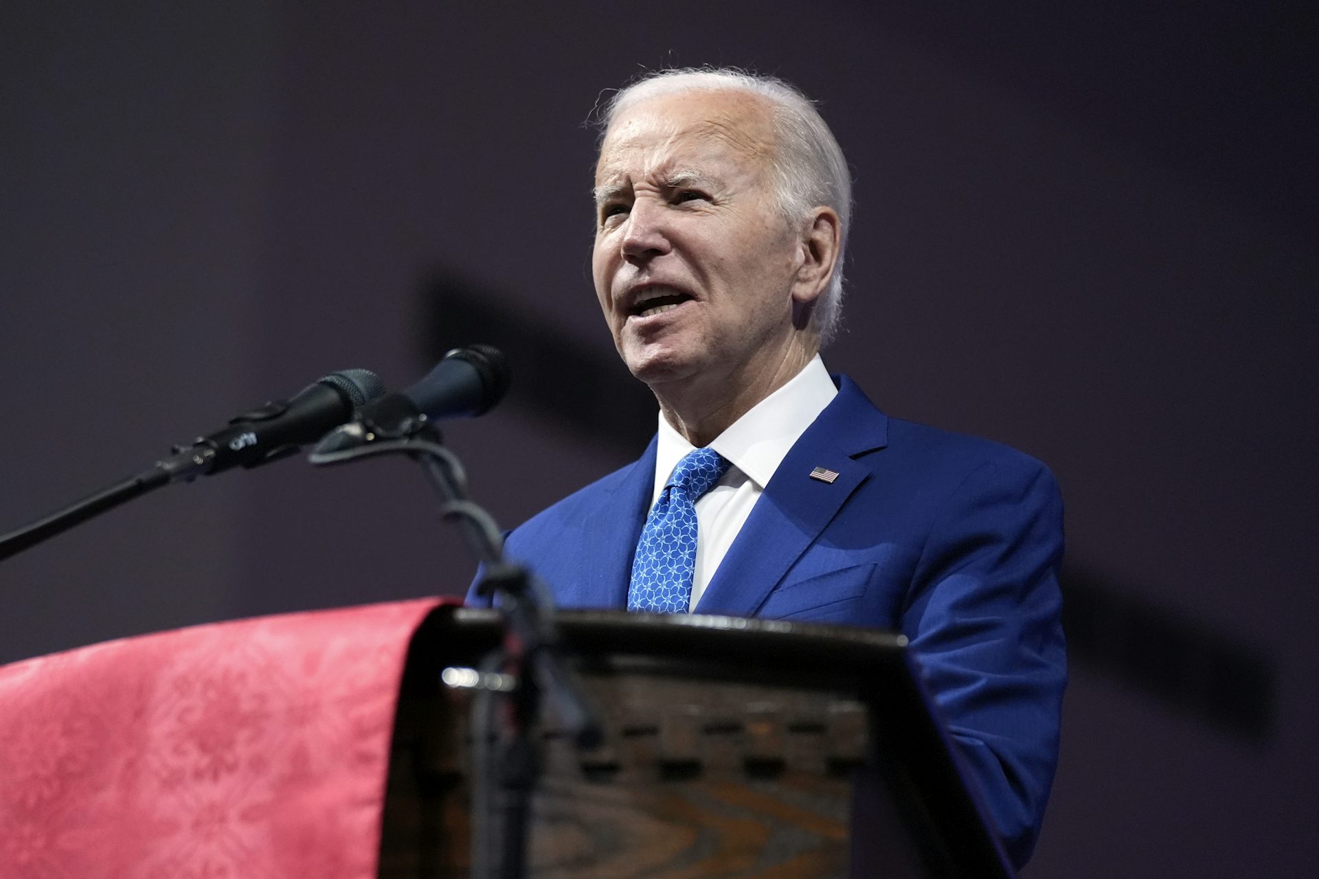 A man with grey hair in a blue suit speaks into a microphone in a church.