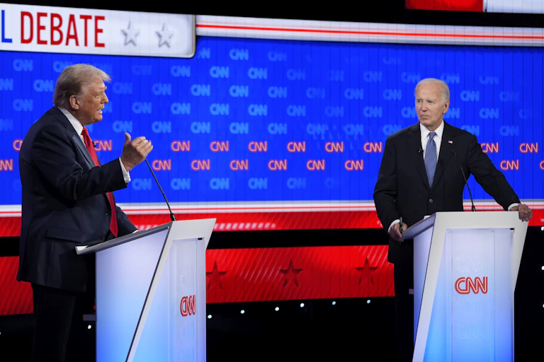 Two men stand behind podiums; one on the left with grey-blond hair gestures as he speaks as the other looks at him.