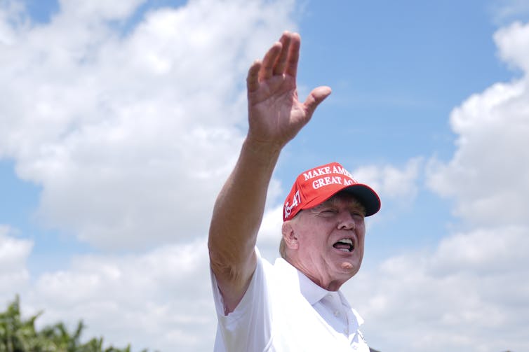 An older man in a white golf shirt and red Make America Great Again ball cap waves.