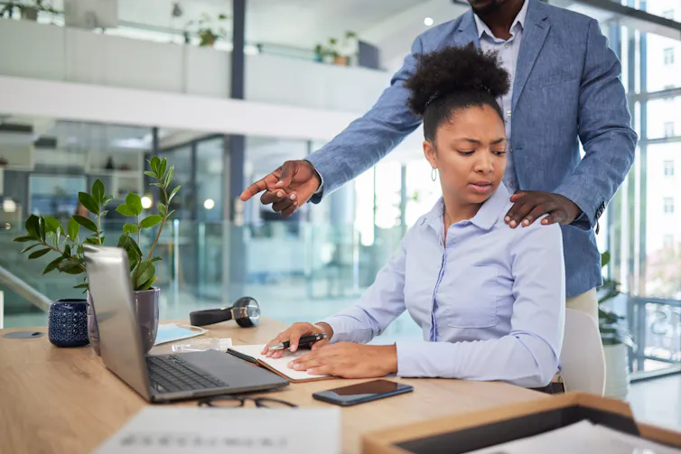 A women sitting a a desk with a concerned look as a man stands with his hand on her shoulder.