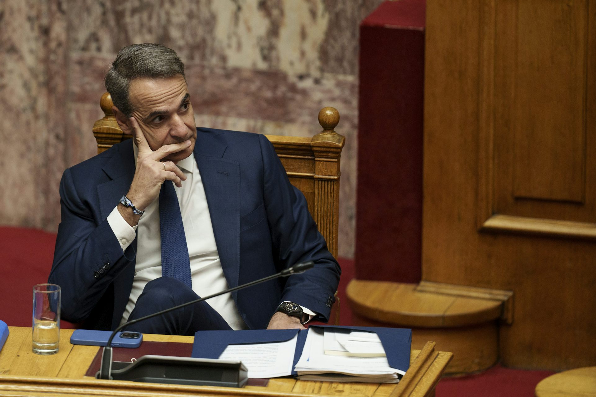 A middle-aged man in a navy suit sits at a desk with his face resting against his hand