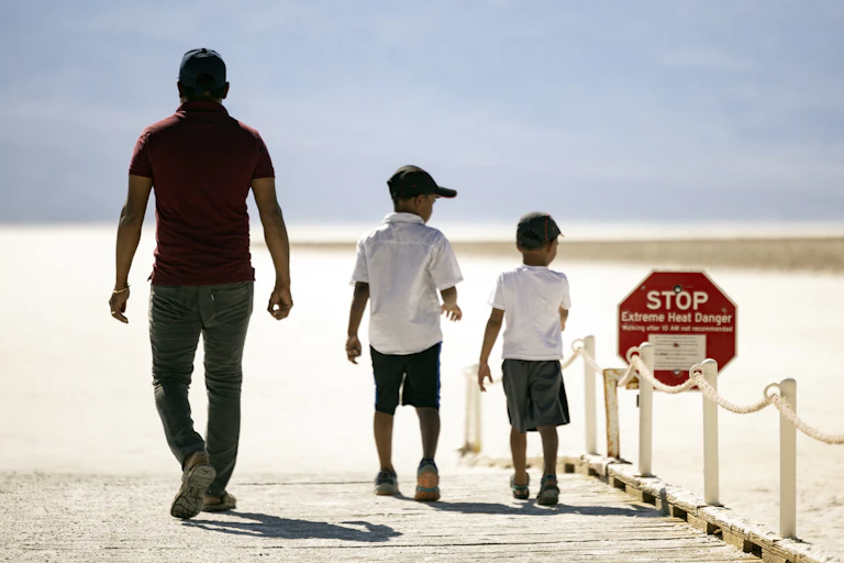 A man and two young boys walk toward a desert landscape, passing a sign that says "Stop: Extreme Heat Danger"