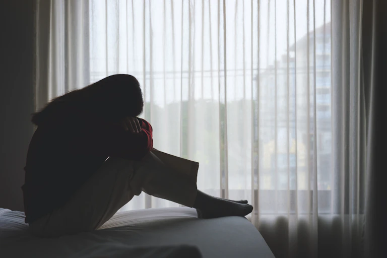 A woman sits on a bed with her head on her folded arms