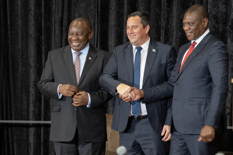 Two men shake hands while standing next to another man. All wearing suits and smiling.