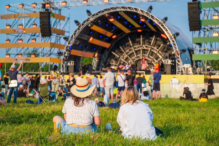 two women sit on the grass in front of a stage at a music festival