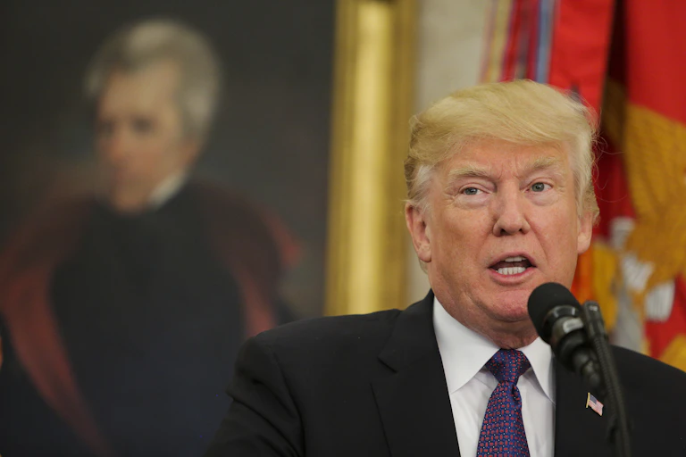 A white man dressed in a business suit stands near a portrait of a former US president.