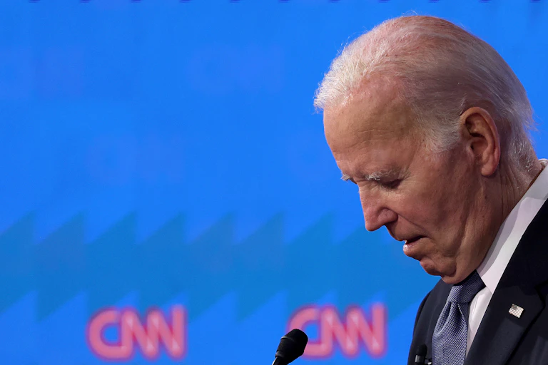 A man in a suit and tie, standing at a microphone with his head bowed.