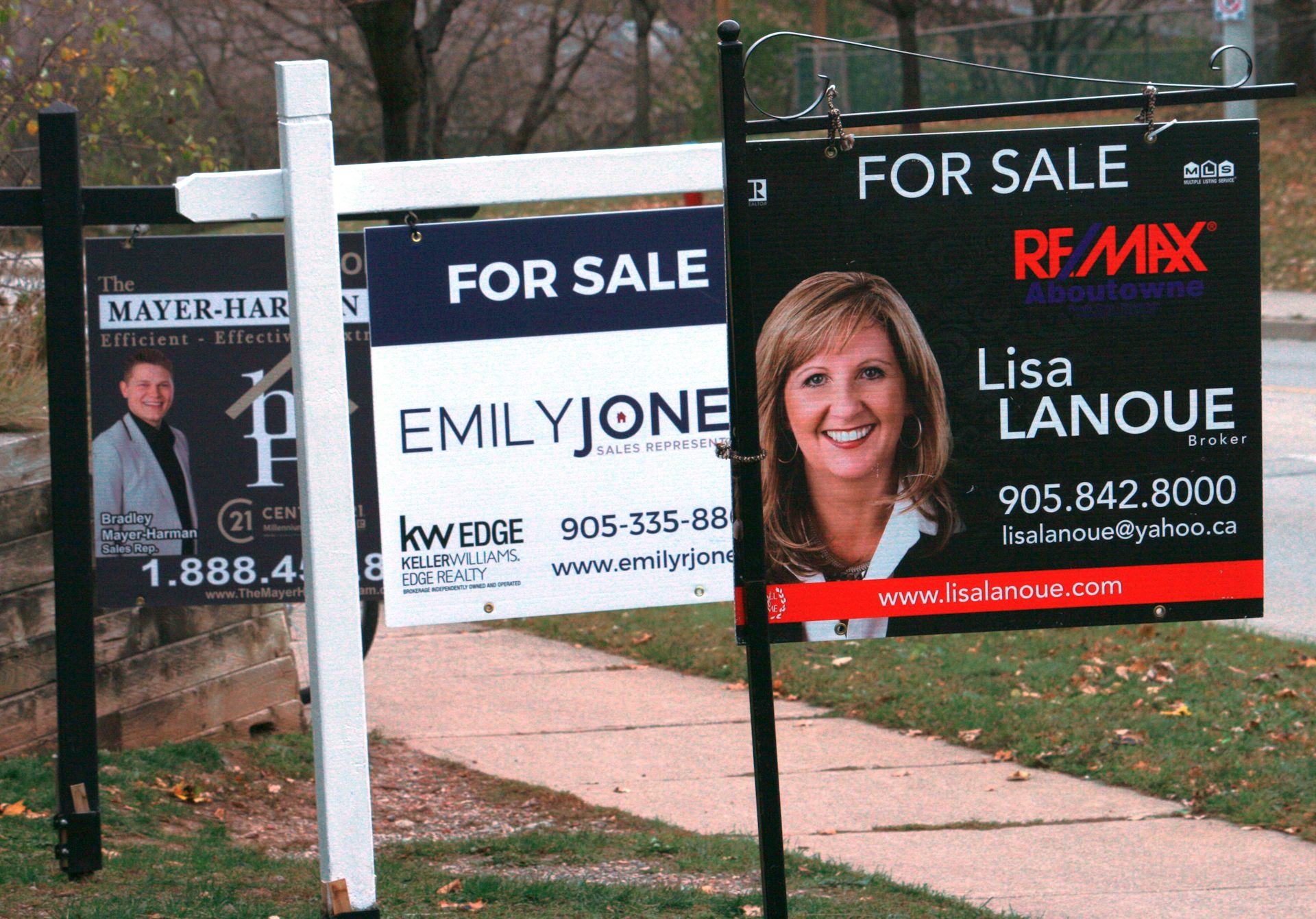 Signs that say 'For Sale' alongside photos and names of real estate agents are seen sticking up out of the ground beside a sidewalk