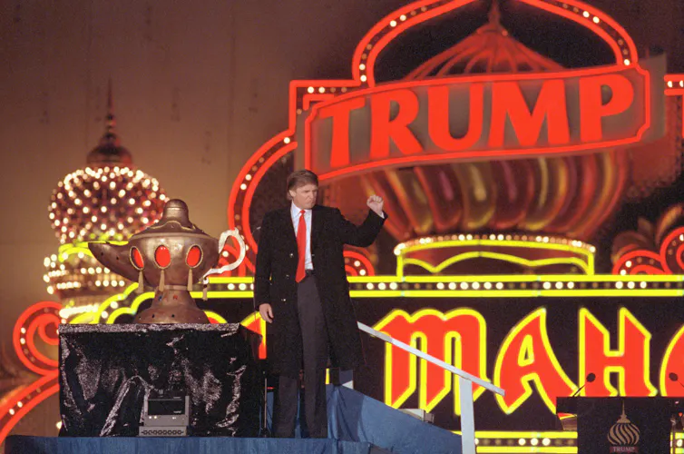 A man in a long dark coat and tie raising his fist in front of a brightly lit building and sign.