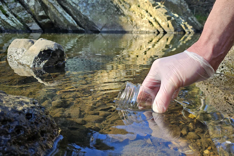 A gloved hand reaches into a waterway to collect a sample in a jar