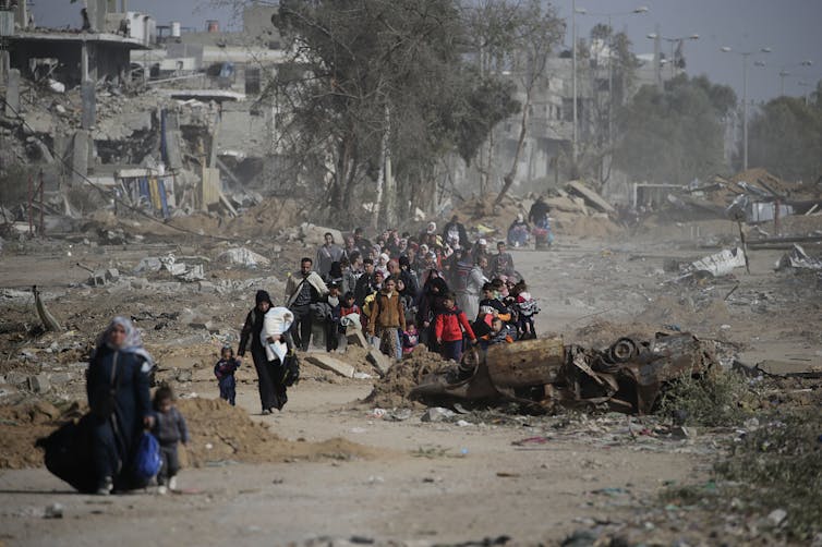 A group of people walk together, carrying their possessions through rubble.