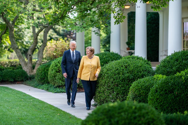 Angela Merkel, wearing a yellow jacket and black trousers, walks with Joe Biden, wearing a navy suit, in the White House rose garden
