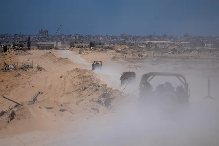 Three Israeli army vehicles driving down a dusty road towards a war-damaged city.