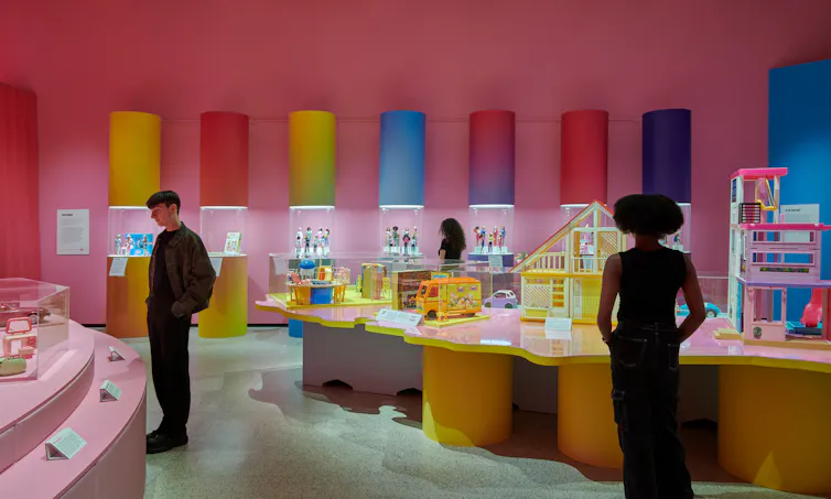 People looking around a pink exhibition filled with Barbie accessories and dolls.