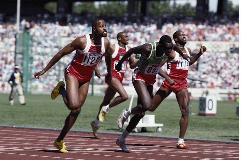 A vintage photo in colour of four men in athletics shorts and vests finishing a race on a track, some grimacing.