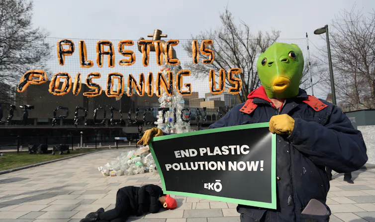 un homme déguisé tient une pancarte et une autre personne est allongée derrière avec un slogan au-dessus contre le plastique