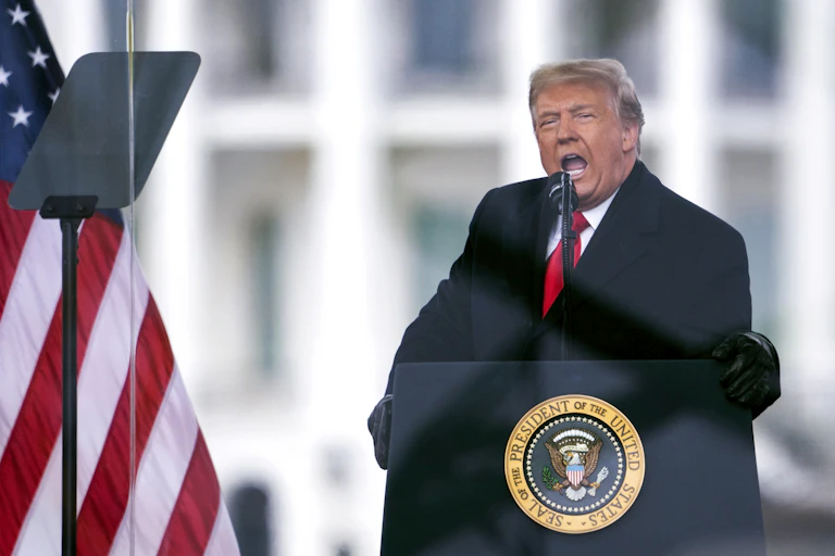 A man dressed in a black suit and red tie, speaking at the lectern, with the American flag next to it.