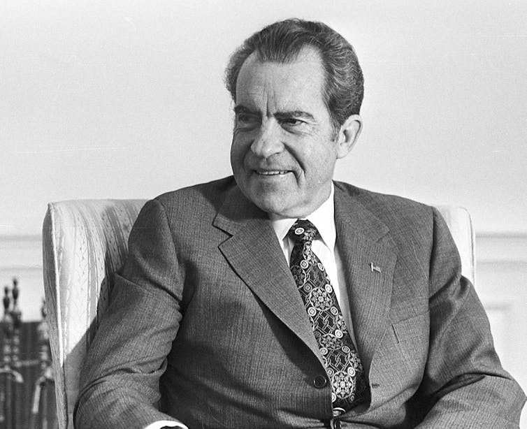 A black and white photo of a man in a black suit and tie, looking sideways and smiling.
