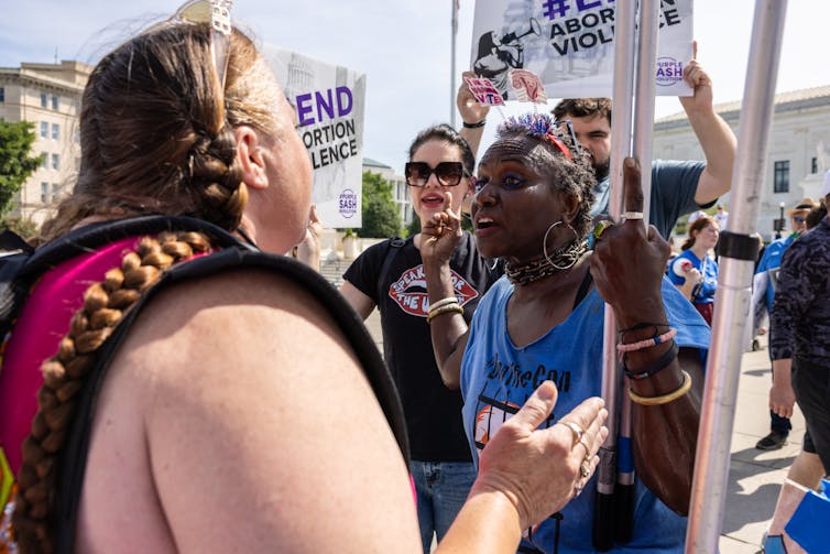Two women face each other. People stand behind the older woman, who is Black, and have sign that says 'End abortion violence.'