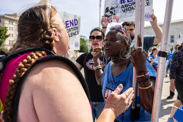 Two women face each other. People stand behind the older woman, who is Black, and have sign that says 'End abortion violence.'